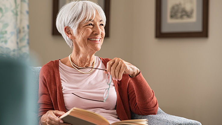 An older woman looks up from reading a book and grins, revealing shiny white teeth