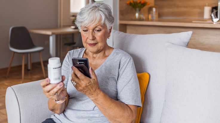 Sitting on her living room couch, an older woman is researching information about her prescription using the bottle in her right hand and smartphone in her left hand.