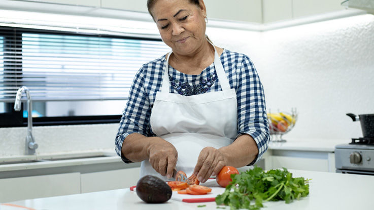 Standing at her kitchen counter, an older woman slices a tomato on a cutting board with sprigs of cilantro and an avocado nearby.