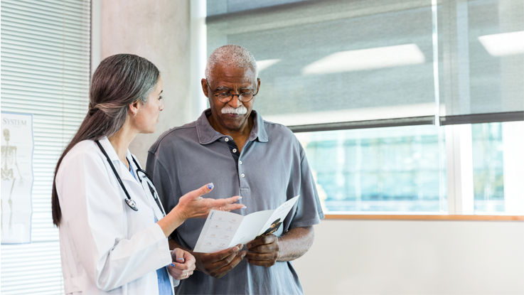 Standing in a clinic waiting room, an older man looks thoughtfully at a pamphlet and listens to his doctor as she gives advice.