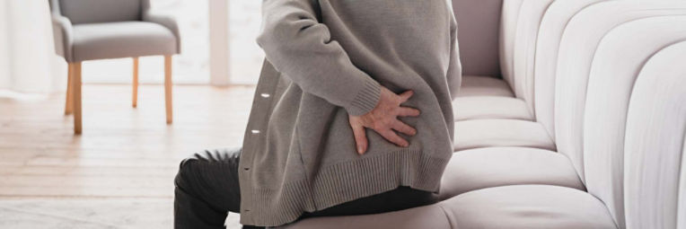 Woman seated on couch holds her hand to her lower back to provide support.