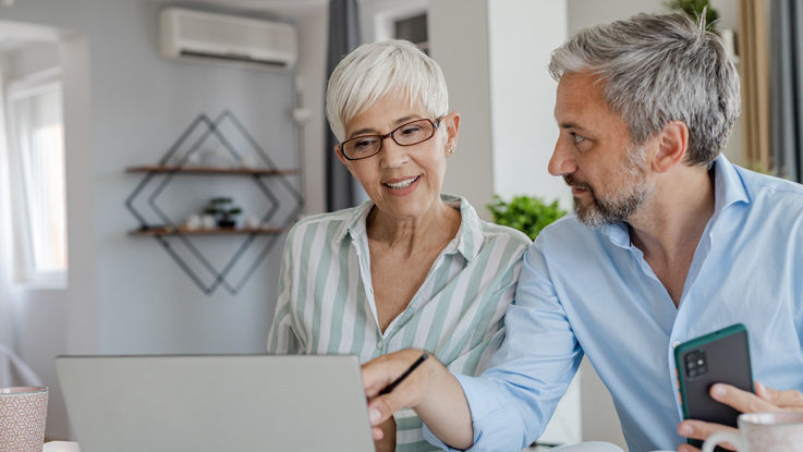 A senior couple sits at their dining room table to shop for Medicare plans on a laptop. 