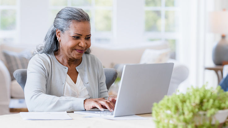 A senior woman sits at her dining room table to compare Medicare plans on her laptop. 