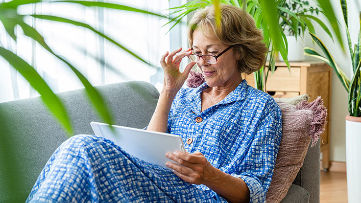 A woman in a blue dress looks at a computer tablet while resting on a couch.