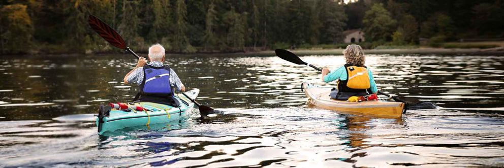 Two older adults paddle kayaks on a calm lake.