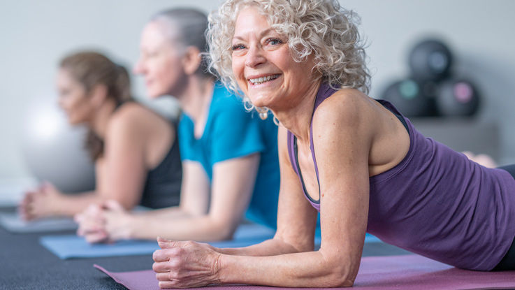 In yoga class, an older woman smiles as she exercises on a yoga mat as other class participants do the same in the background