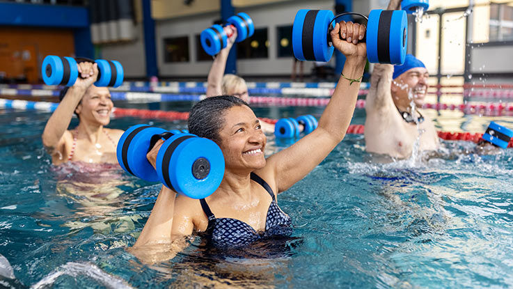 A group of adults lifting foam dumbbells above their heads while standing in a pool.