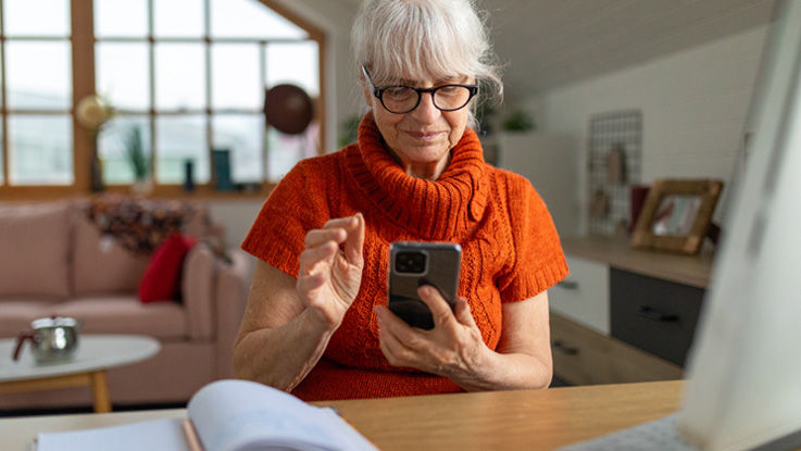 Sitting at her living room desk, an older woman shops on her smartphone.