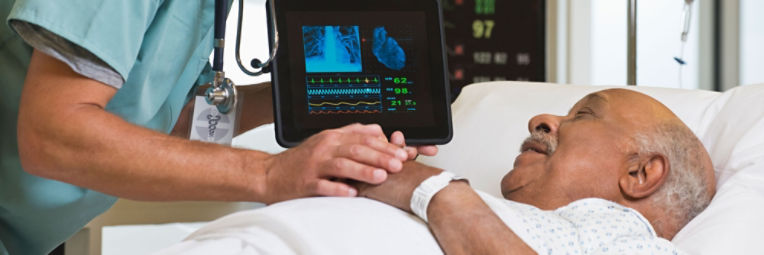 A doctor rests a hand on the hands of an older patient in a hospital bed as he explains the results of a heart scan.