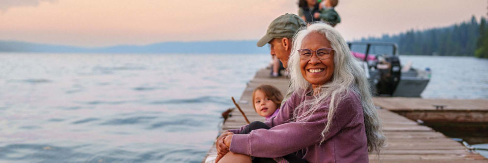 An older woman smiles as she sits on a lake doc at sunset with her husband and grandchild