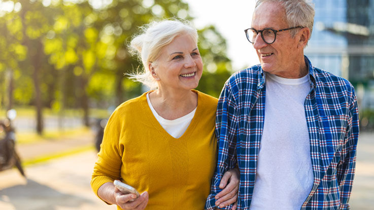 An older husband and wife walk down a city street on a sunny summer day, shoulder-to-shoulder as the wife holds her husband’s arm.