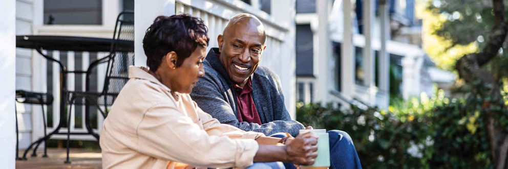 An older woman and older man drink their morning coffee on their front porch