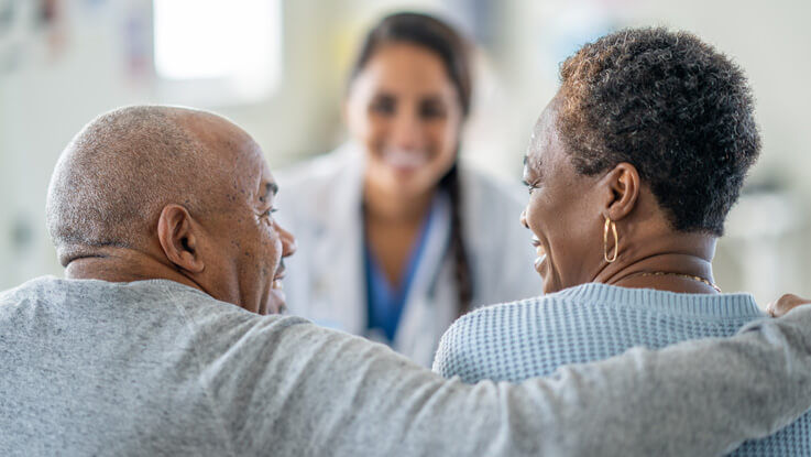 Sitting across from a doctor, an older married couple smile at each other as the husband hugs his wife’s shoulders.