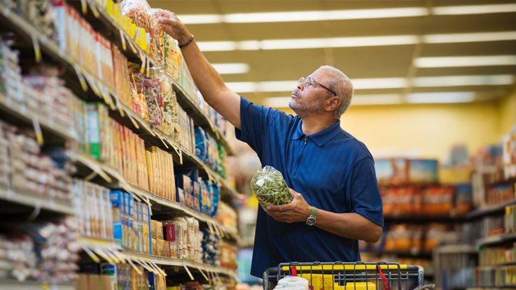Older man walking through a grocery store, picking items from a shelf and putting them in his cart.