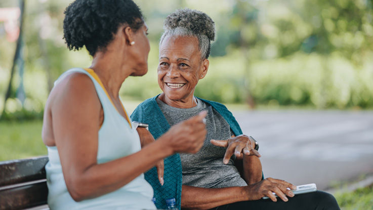 Sitting on a park bench after a workout, a woman smiles and gestures as she talks to an older woman.