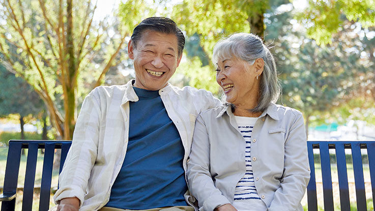 An older couple sits on a park bench, smiling.