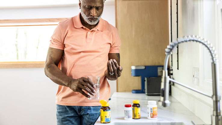 Standing at his kitchen sink, an older man is ready to a prescription pill as he holds a glass of water.