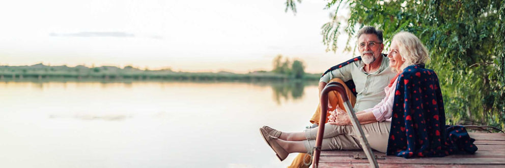 A couple sits together on a dock. They share a blanket wrapped around their shoulders and look out at the lake as the sun sets.