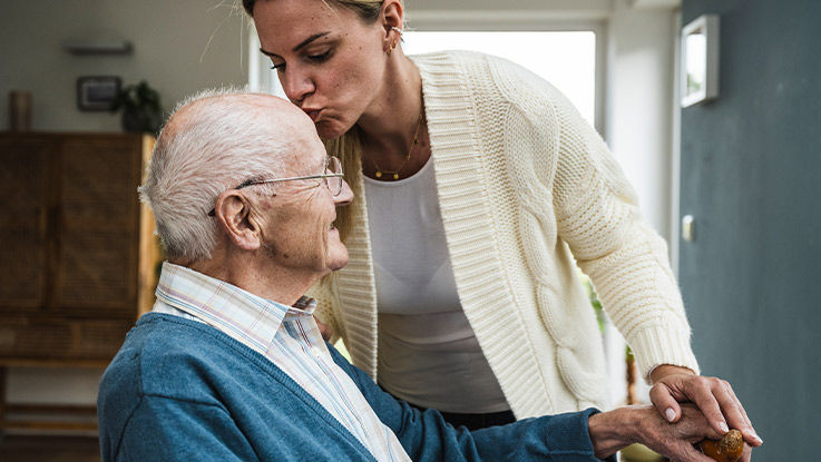 Caregiver leaning down and kissing the forehead of an older man sitting in his home.