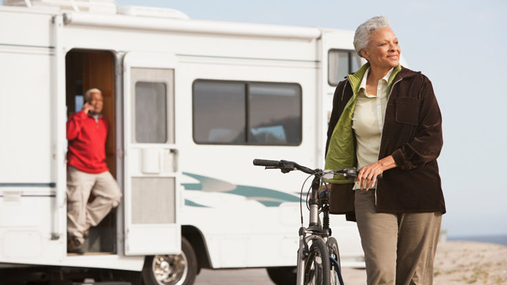 Older woman standing and holding a bicycle in the foreground, and an older man in the background standing in the doorway of an RV, talking on the phone.