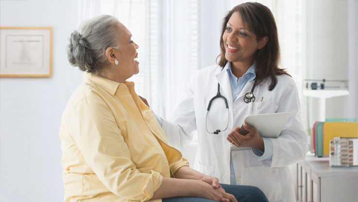 In her exam room, a female doctor places her hand on her older female patient’s shoulder, smiling as her seated patient laughs.