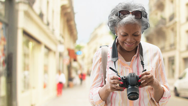 Older woman standing outside, looking at the screen of a digital camera that's around her neck.