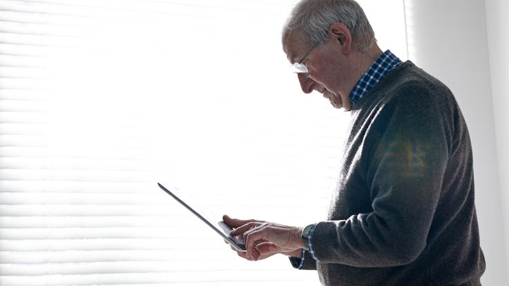 Standing in front of his sunlit living room window, an older man browses the screen of the tablet device in his hand.