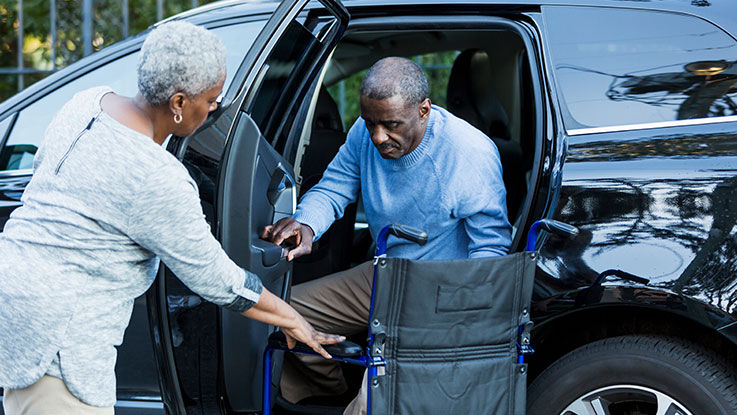 An older woman holds a wheelchair steady for her husband as he gets out of the backseat of a car.