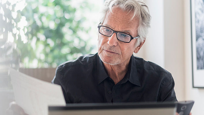 An older man sitting in front of his computer consults information from a piece of paper