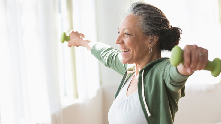 Standing in front of her living room window, an older woman smiles as she works out with dumbbell hand weights.