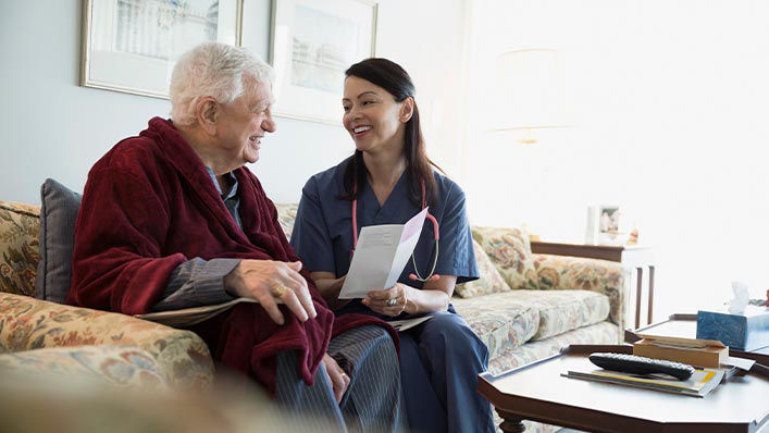 A smiling hospice nurse shares a pamphlet with an elderly man on the couch in his home.