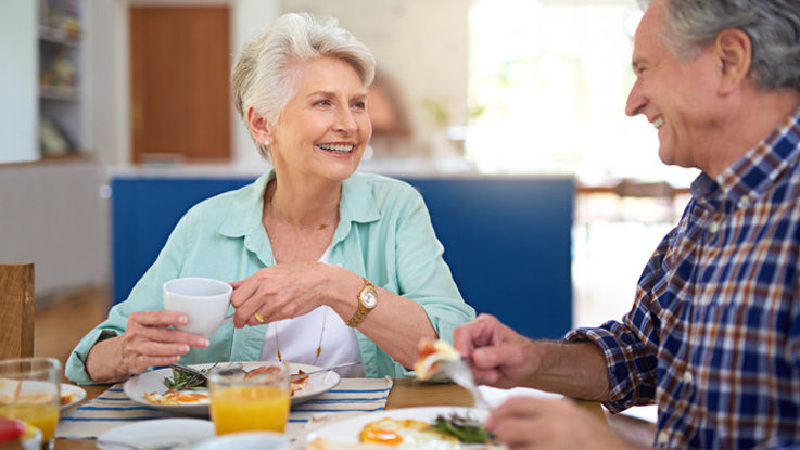 Sitting at their dining room table, an older woman holds a cup of coffee while smiling at her laughing older husband as they both eat breakfast.