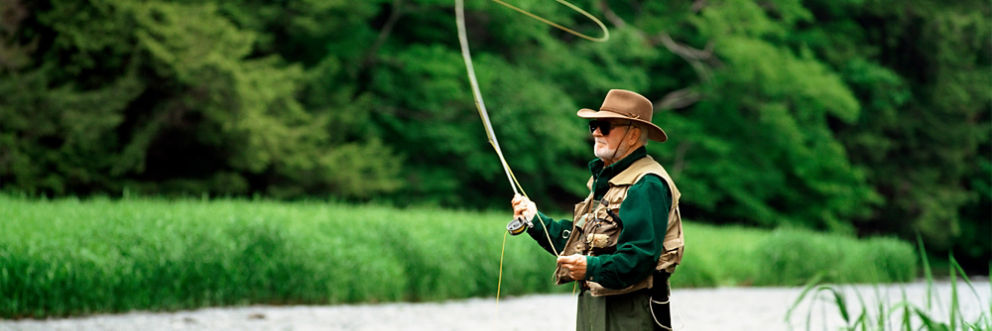Wearing a hat, vest and waders, an older man stands in a river as he fly fishes on a summer day.
