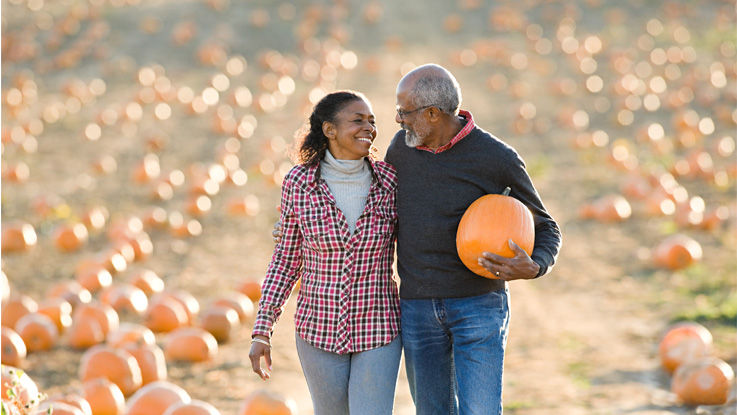 Walking through a pumpkin patch on an autumn day, an older man and woman smile at each other with arms across each other’s backs as the man carries a pumpkin.