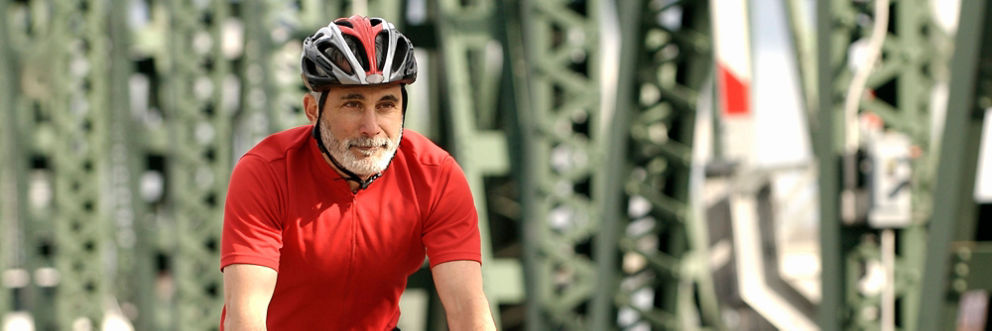 An older gentleman smiles while biking across a bridge.