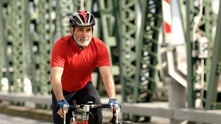 An older male cyclist riding his bike across the bridge with a bike helmet and gloves.