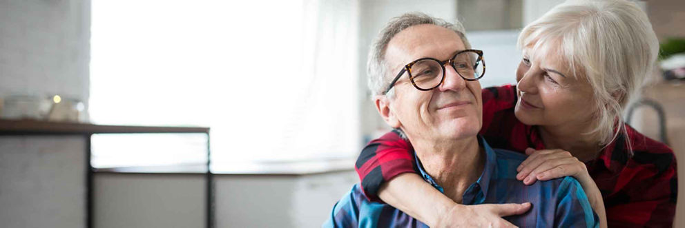 An older man wearing glasses smiles as an older woman hugs him from over his shoulder