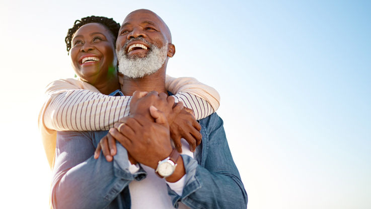 Older woman standing behind older man with her arms around his neck, smiling and looking into the distance.