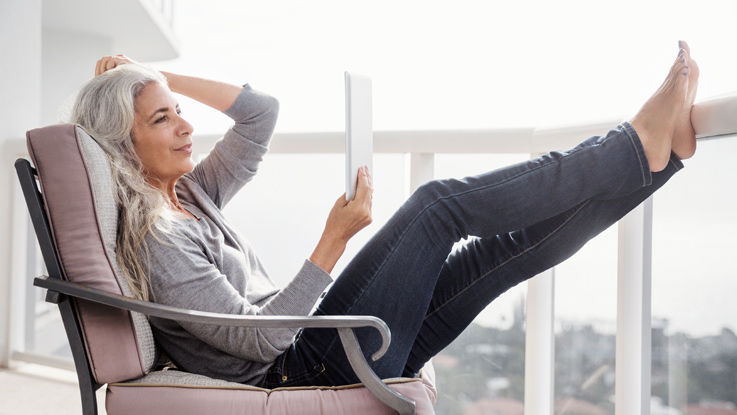 A senior woman lounges in a chair on a balcony while comparing Medicare plans on her tablet. 