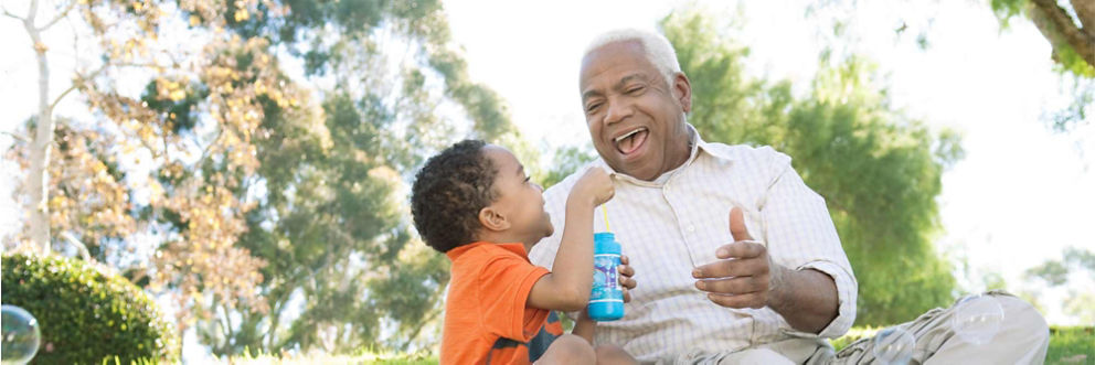 An older man sits with his grandson at the park as they laugh and blow bubbles together.