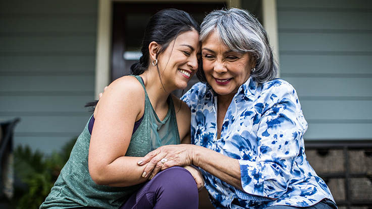 An older woman sits on the steps of a porch with her daughter, touching their heads together and smiling.