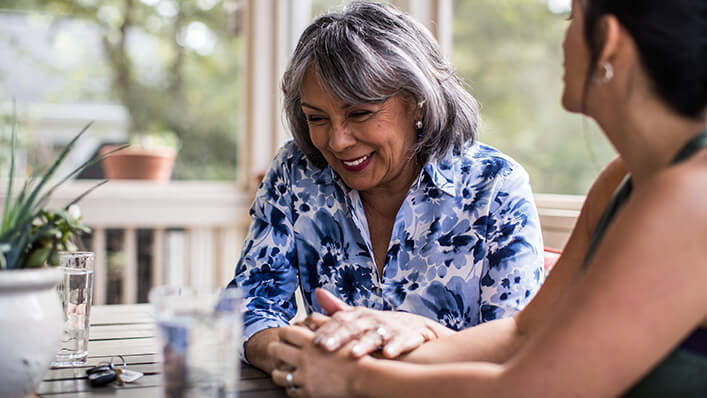 A woman and her laughing mother sit at a table on the porch and hold hands.