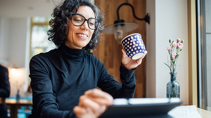 A woman sits at her dining room table smiling as she scrolls through information on her tablet