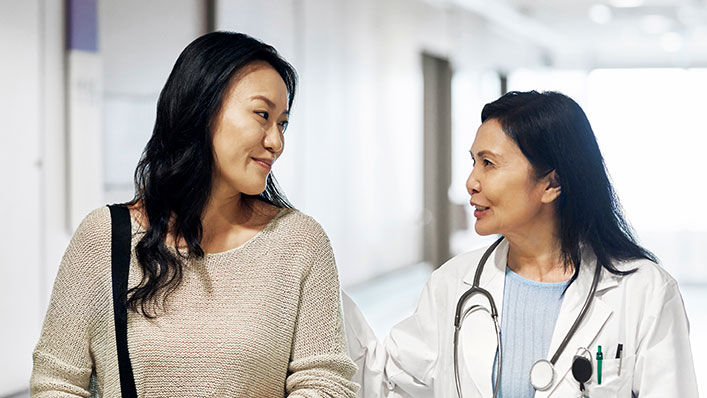 Smiling mature doctor talking to woman in hospital. Female patient visiting healthcare worker for routine checkup. They are standing in corridor.