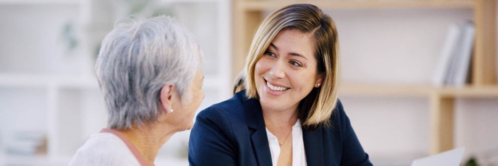 Sitting on a living room couch, a female broker is smiling while talking with a smiling older woman.