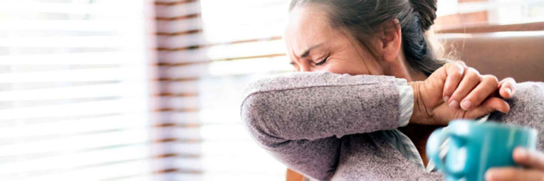 A sick woman coughs into her inner elbow while holding a cup of tea.