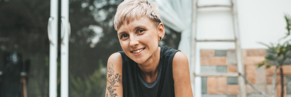 A person with short hair and an arm tattoo smiles on their patio.