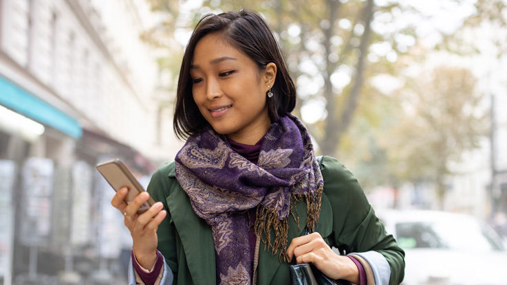 Woman walking down a street looking at the phone in her hand and smiling.