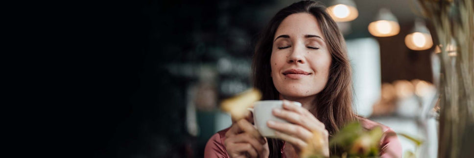 A woman with brown hair closes her eyes as she smells a cup of coffee.