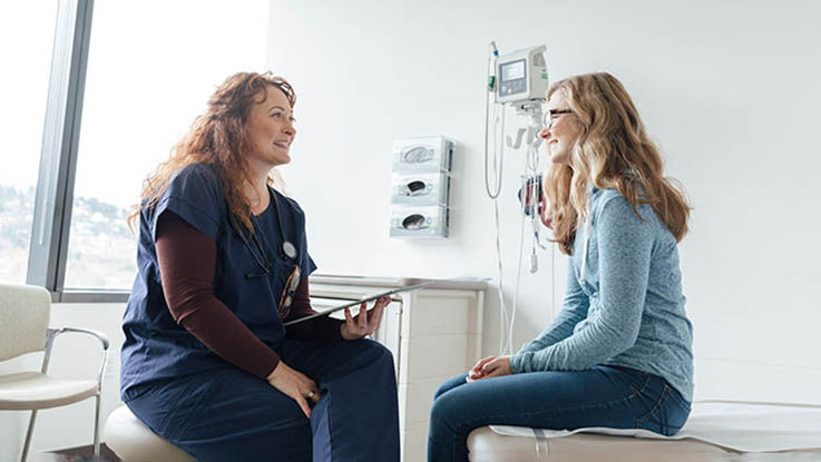 A female nurse chats with a female patient in the exam room to add information to her chart. 